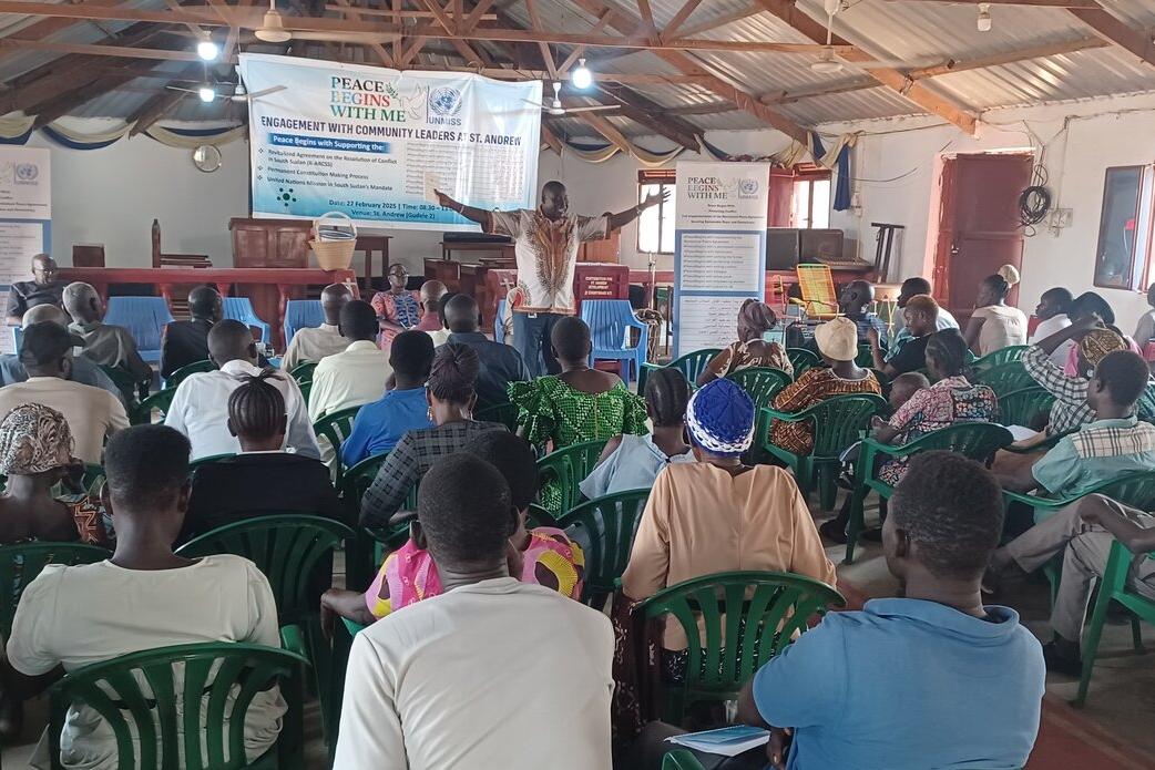 A group of people seated in green plastic chairs seated facing a stage where a man is presenting with their arms outstretched.